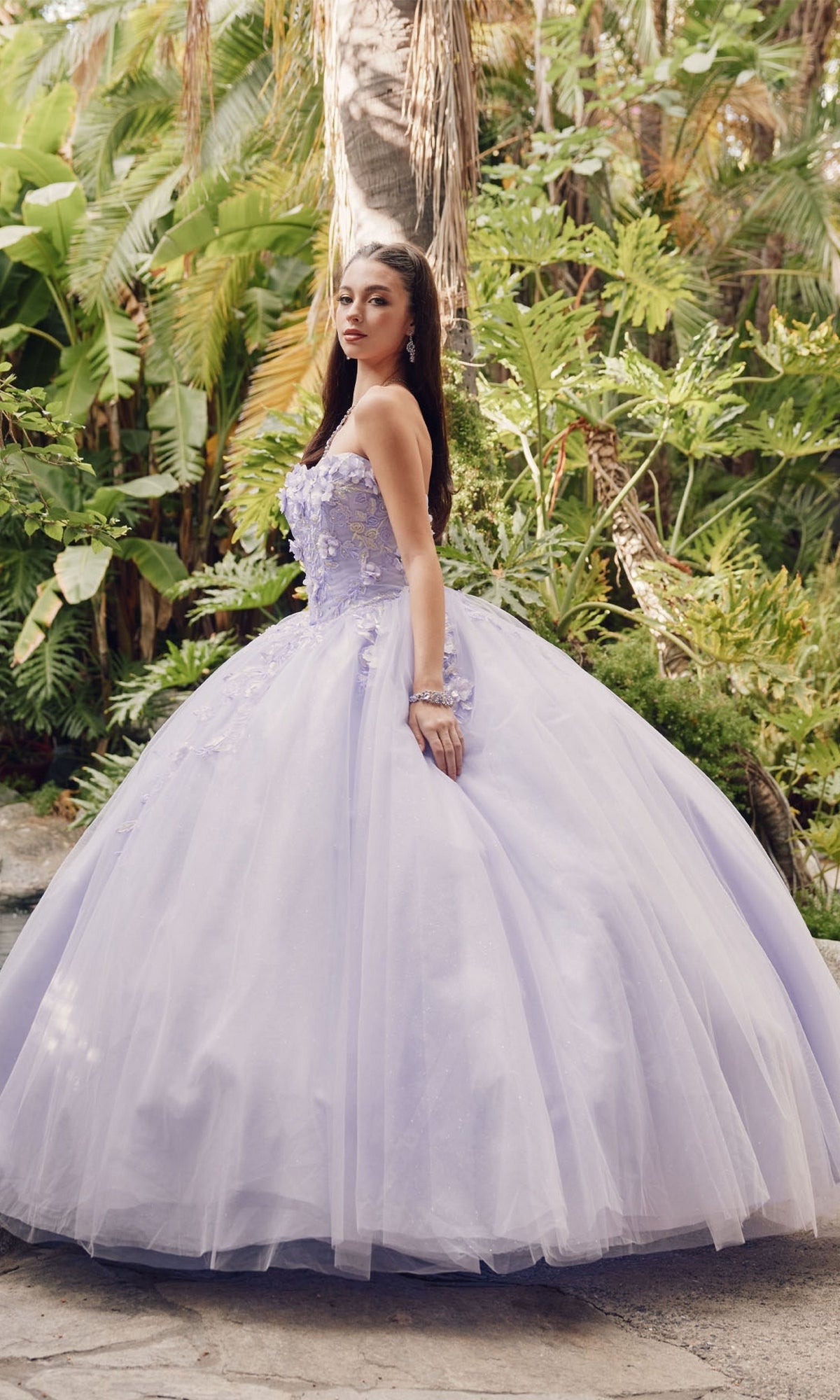 A young woman stands outdoors before lush greenery, wearing the Juliet 1435 Strapless Ball Gown Quinceañera Dress with a full tulle skirt and delicate floral embellishments on the bodice.